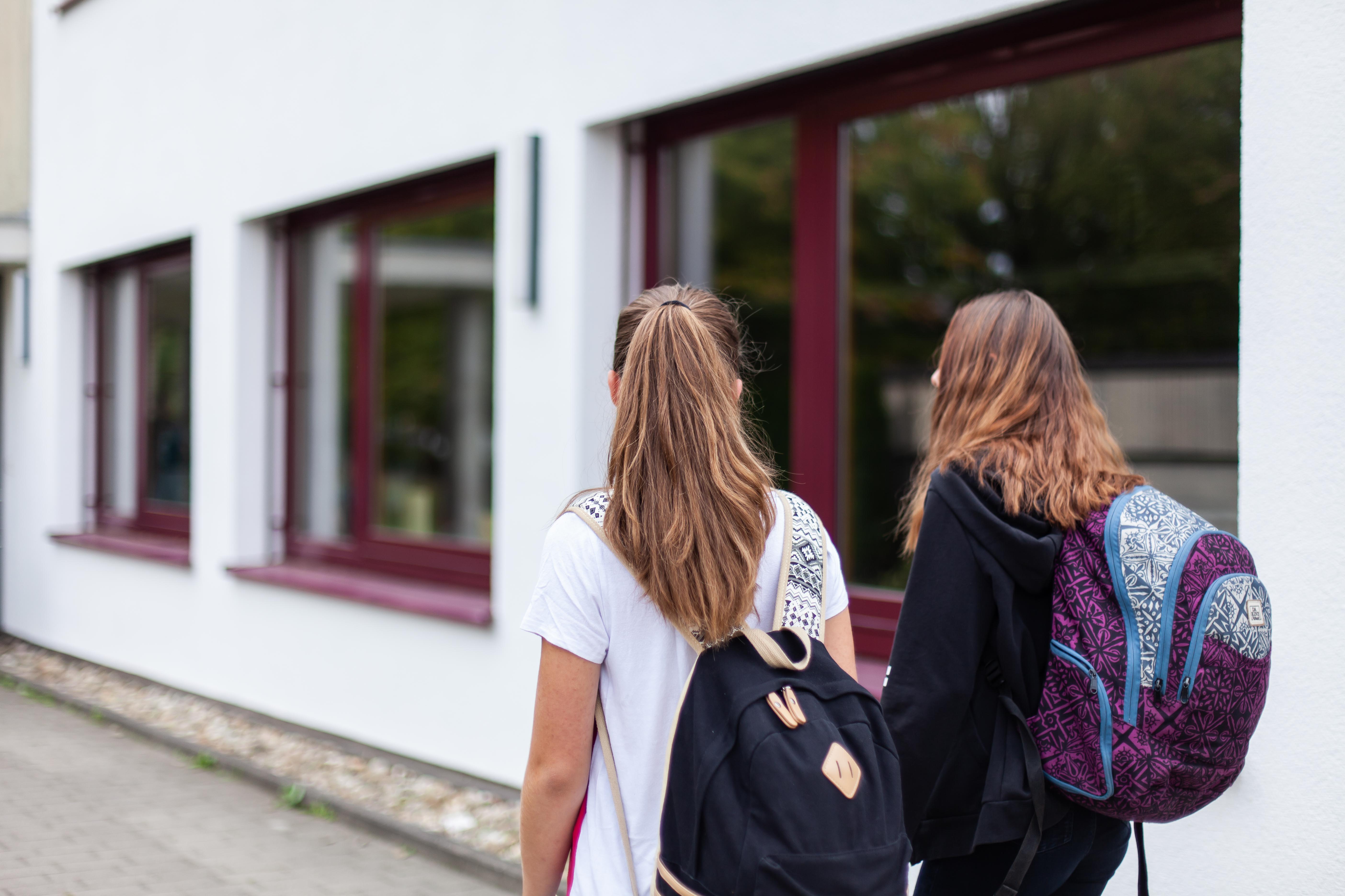 Zwei Schülerinnen laufen nebeneinander auf dem Gehweg neben dem Schulgebäude
