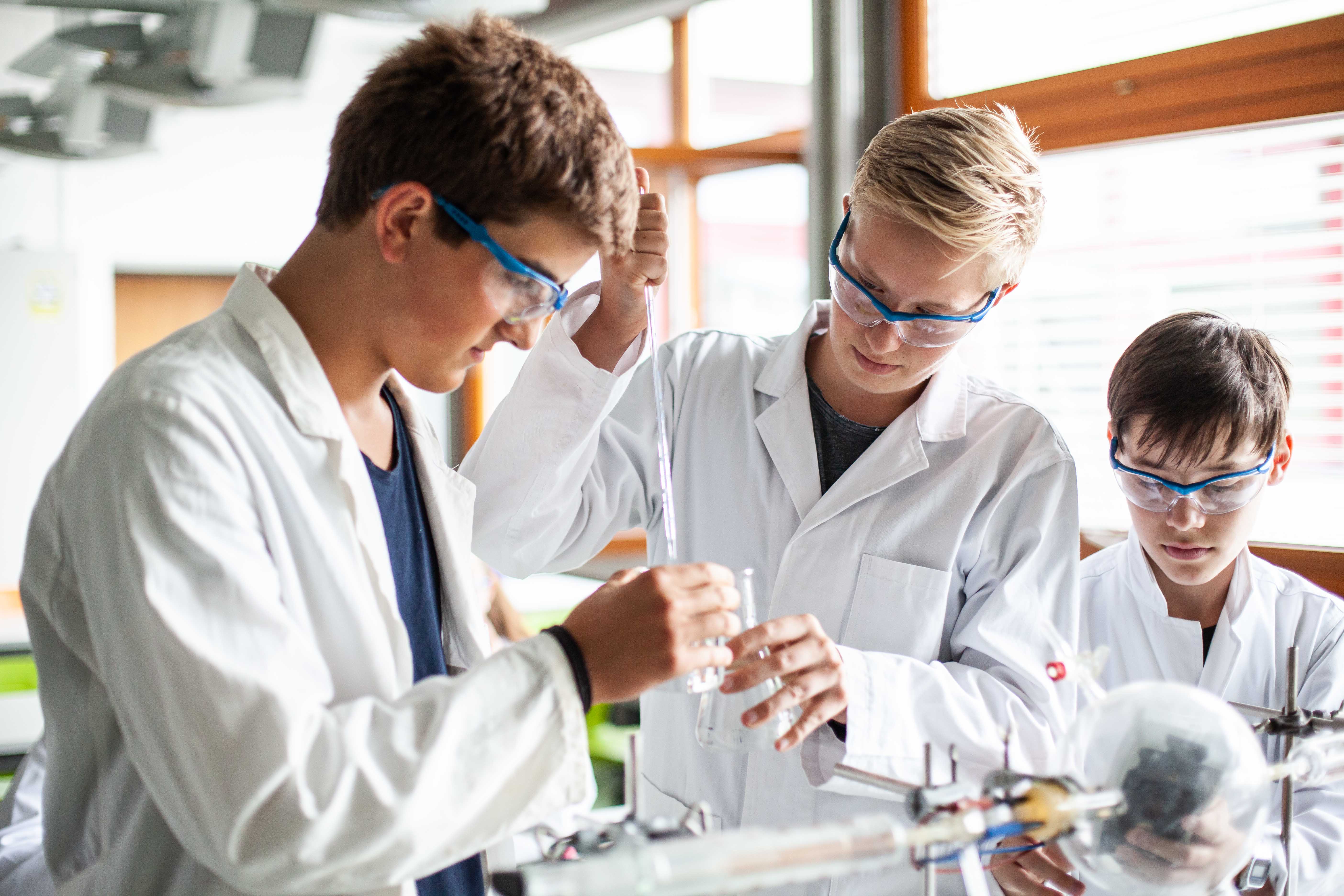 Drei Schüler in Laborkleidung und mit Schutzbrille experimentieren im Chemiesaal.