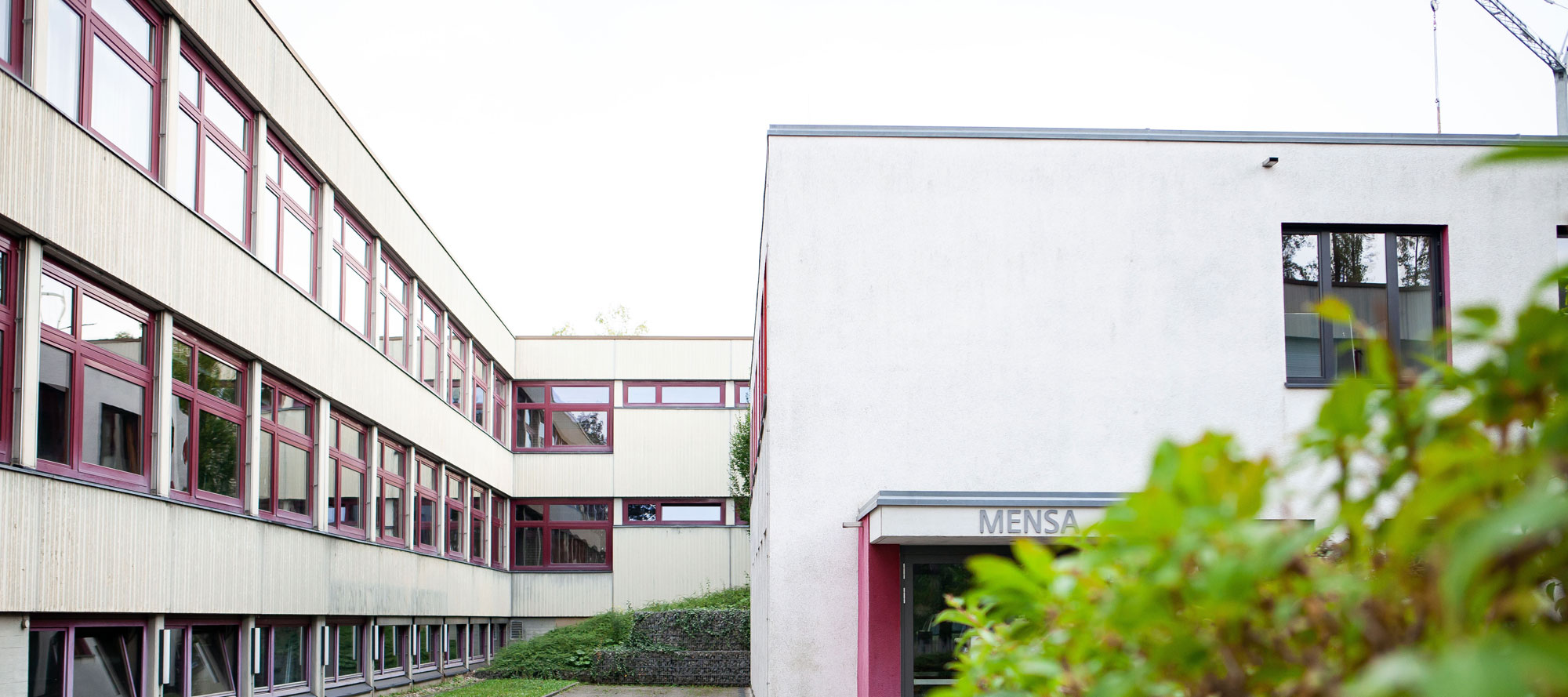 Schulgebäude mit weißer Fassade und roten Fenstern. Über dem Eingang steht "Mensa".