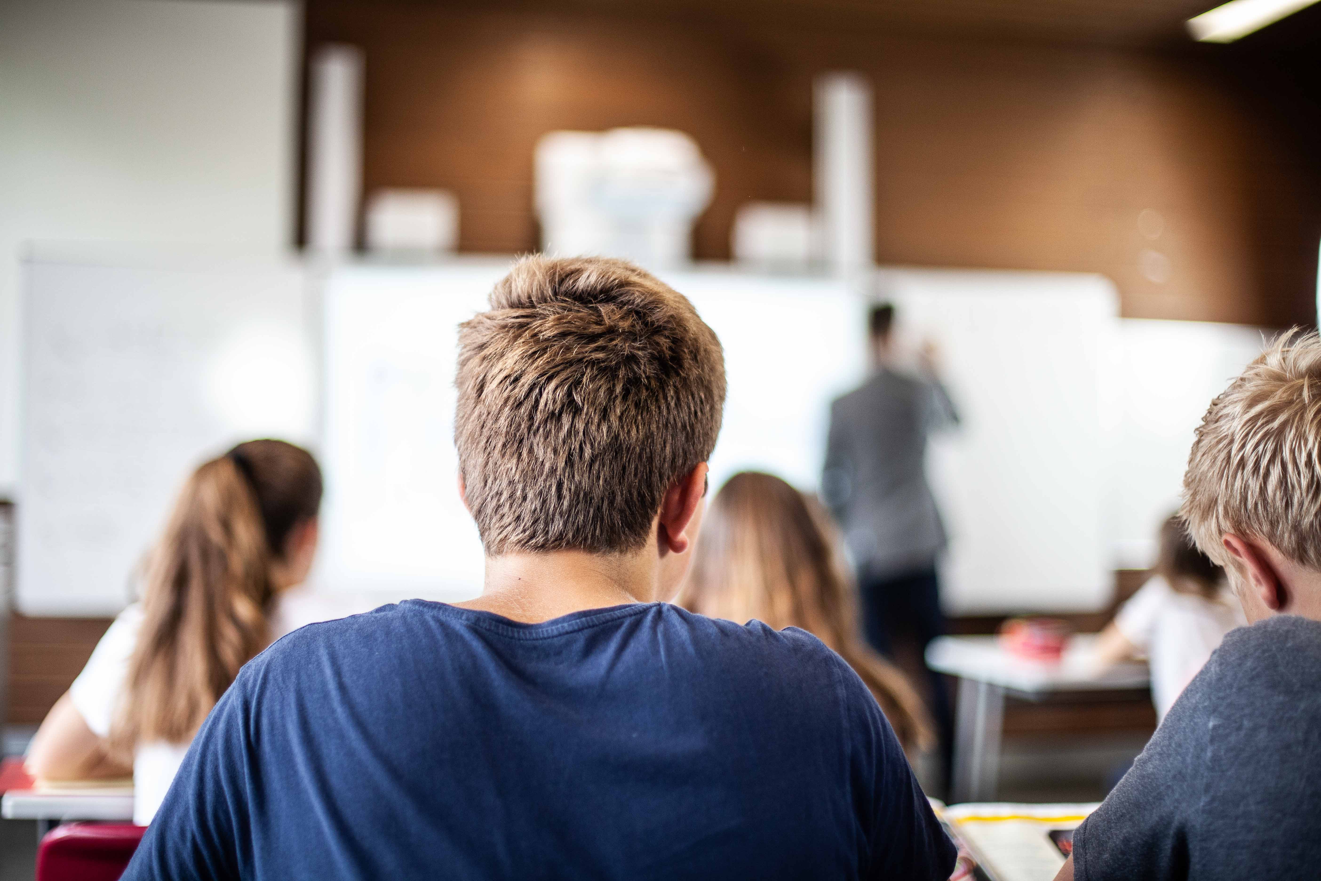 Blick zur Tafel: Eine Klasse im Klassenraum. Der Lehrer steht an der Tafel und schreibt.