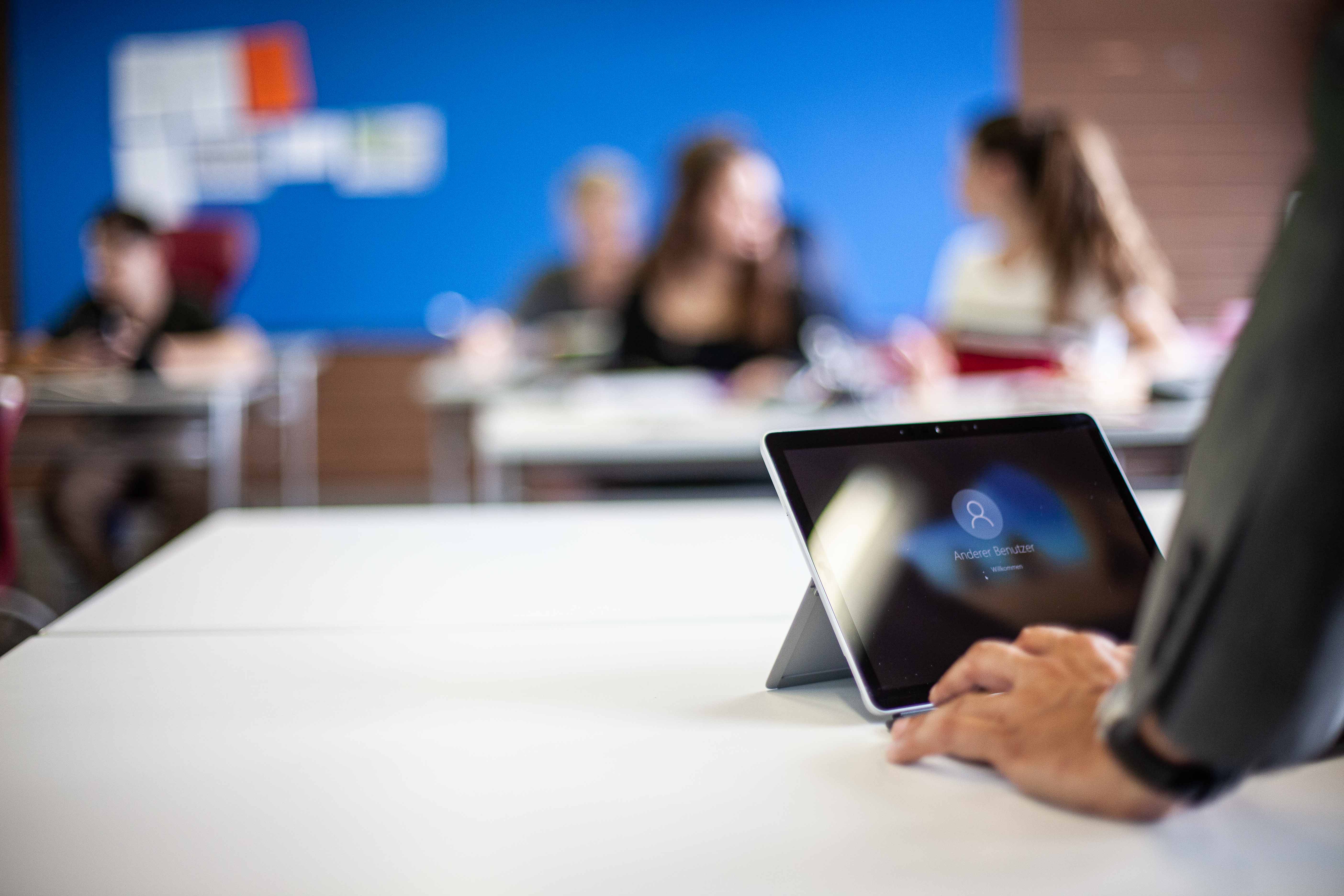 Lehrer am Tablet. Im Hintergrund die Klasse im Klassenraum.