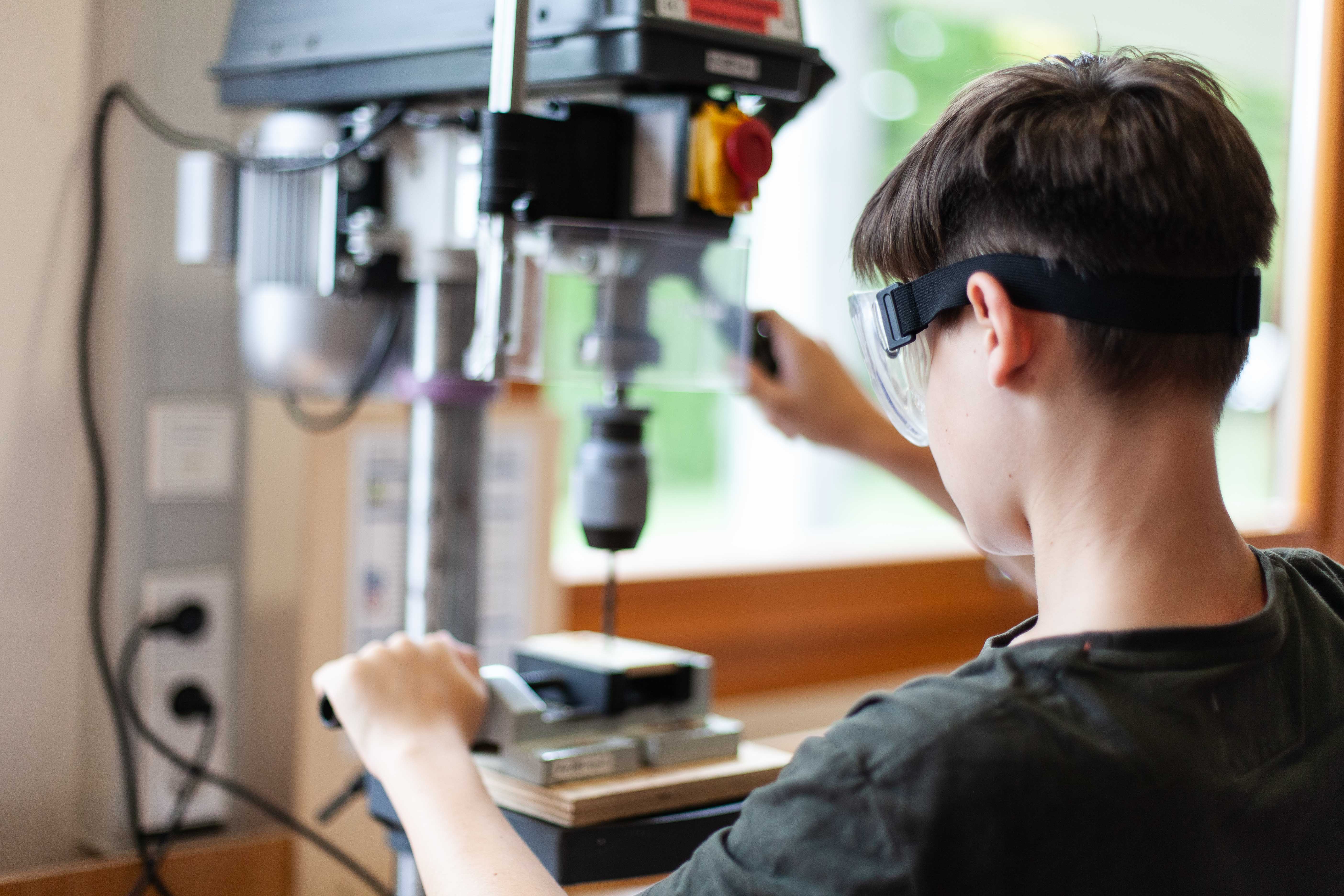 Schüler mit Schutzbrille steht an der Bohrmaschine und bohrt ein Loch in sein Werkstück.