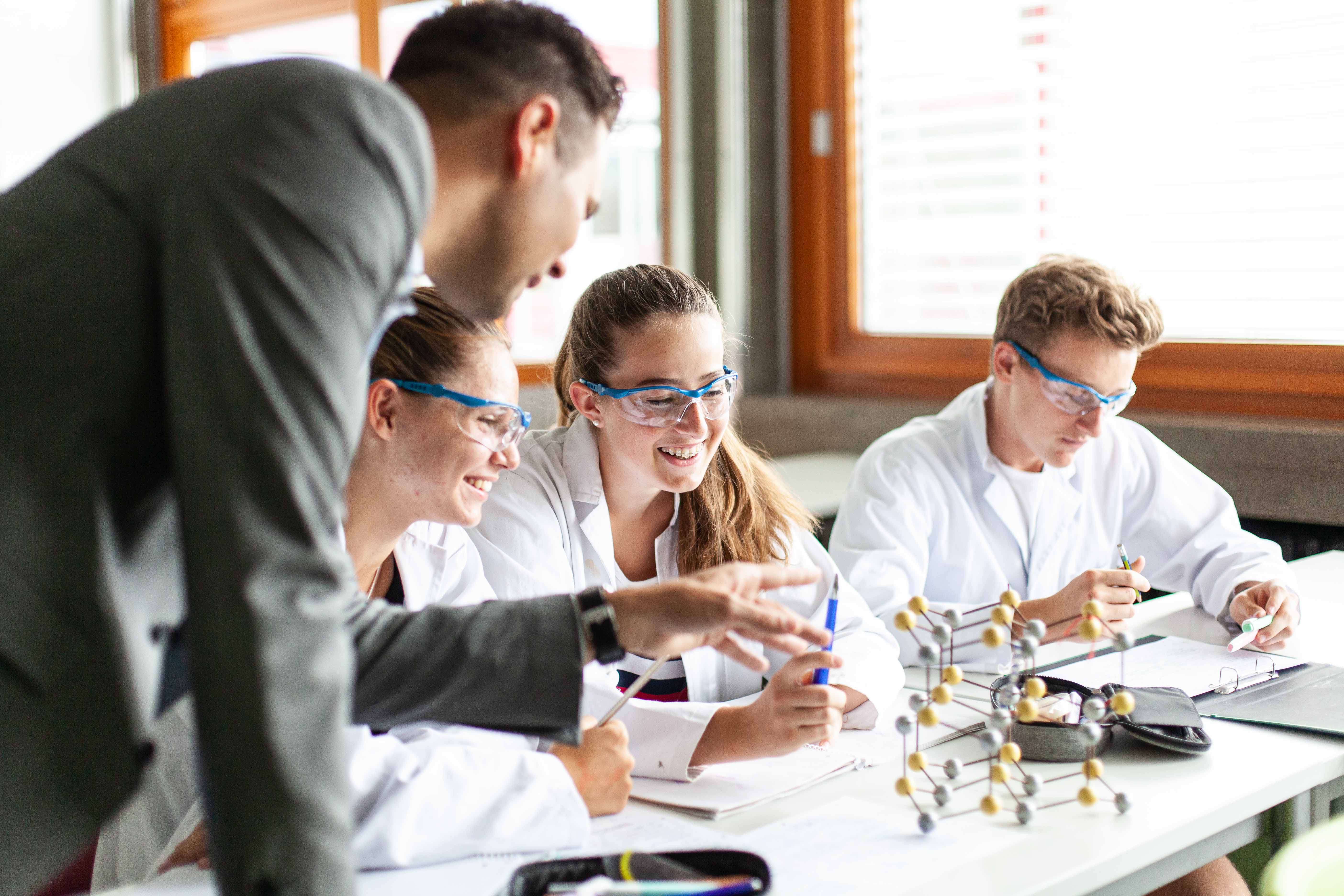 Der Lehrer erklärt den Schülerinnen, welche Laborkleidung anhaben ein Atommodell