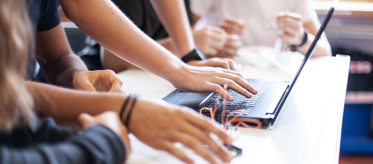Schülerinnen und Schüler sitzen im Physikunterricht um einen Laptop und führen einen Versuch mit elektronischen Schaltkreisen durch.